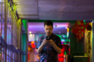 Hombre joven utilizando su teléfono móvil en un entorno urbano iluminado con luces de colores. Representa un enfoque reflexivo en la conexión digital, ideal para ilustrar el concepto de marketing consciente, donde la tecnología y la conciencia se encuentran para crear interacciones más auténticas y significativas.